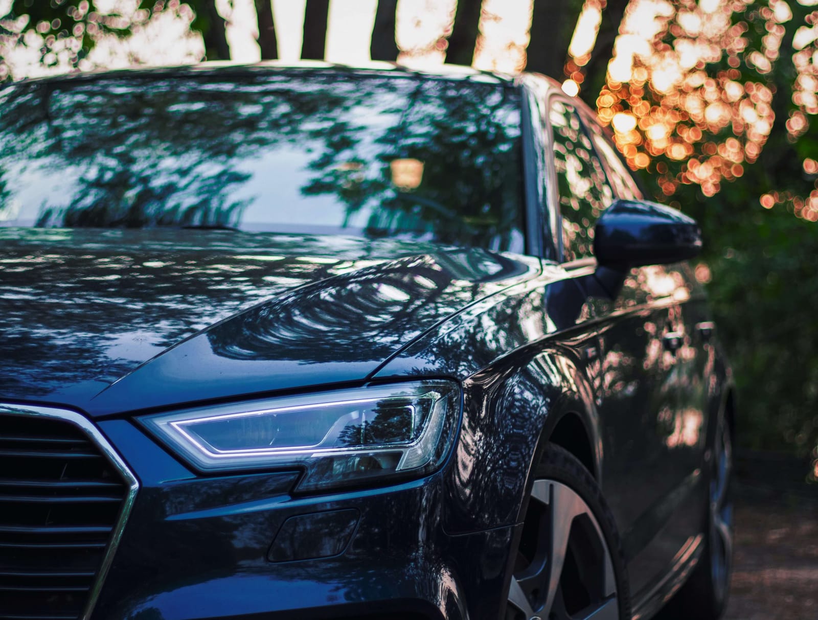 Close-up of the front of a dark blue Audi sedan, its LED headlight lit and trees reflected in the glossy paintwork