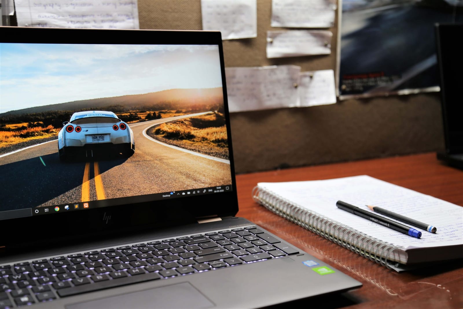 Laptop on a desk showing a white sports car on a winding country road, next to a spiral notebook with a pen and pencil